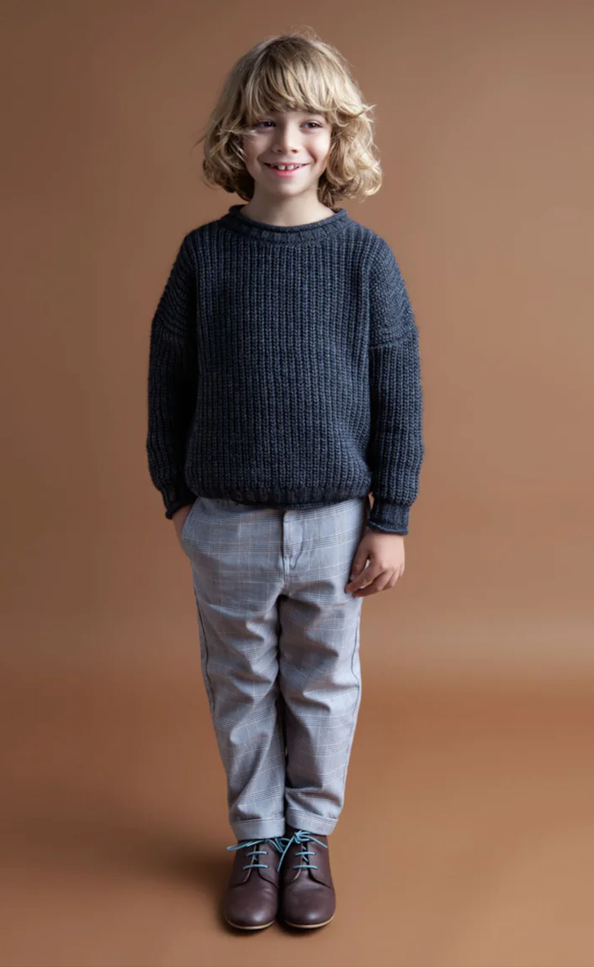 A child wearing light grey plaid trousers and a black sweater with a striped pattern, standing against a brown background.